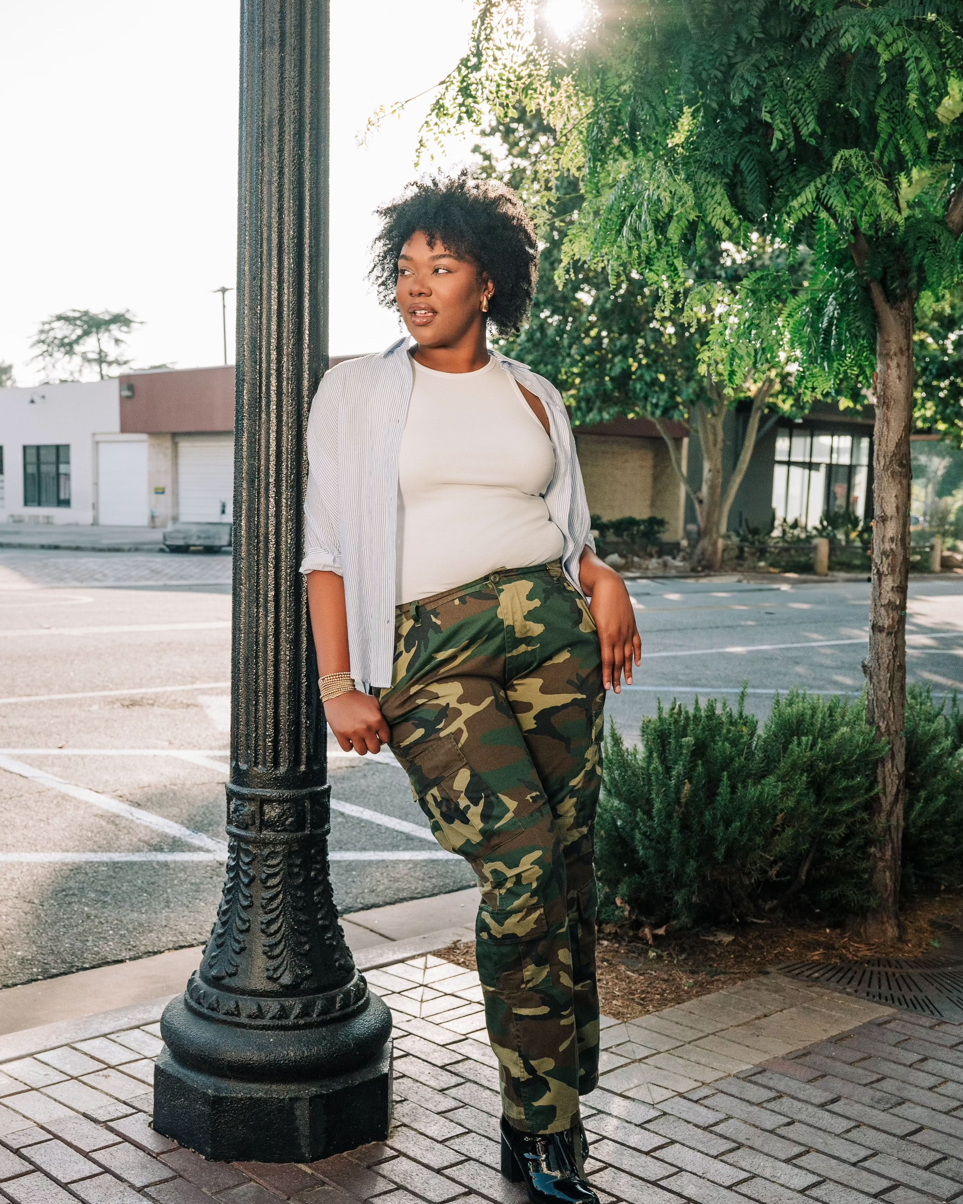 Woman wearing a white ribbed high neck tank top with a white and blue vertical button down with camo pants and black shoes.
