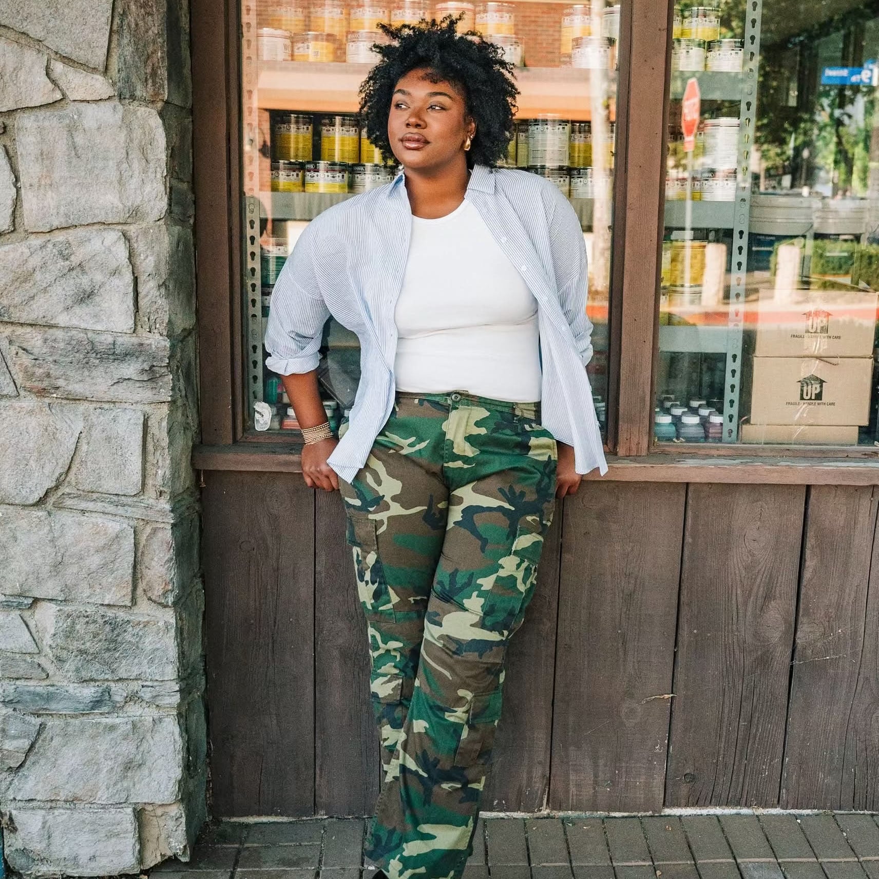 Woman wearing a white ribbed high neck tank top with a white and blue vertical button down with camo pants.
