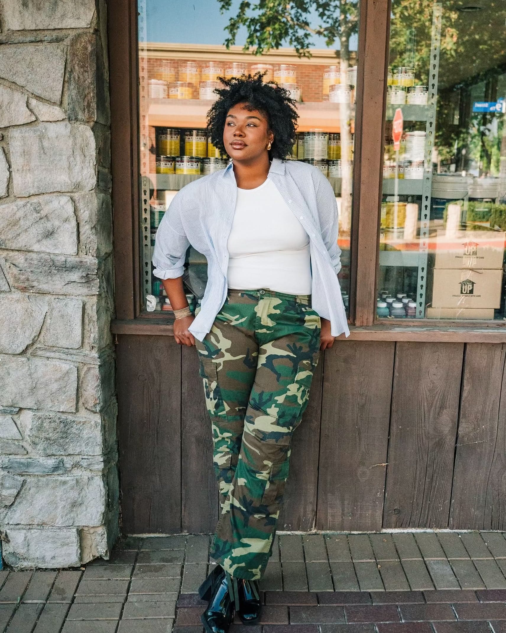 Woman wearing a white ribbed high neck tank top with a white and blue vertical button down with camo pants.