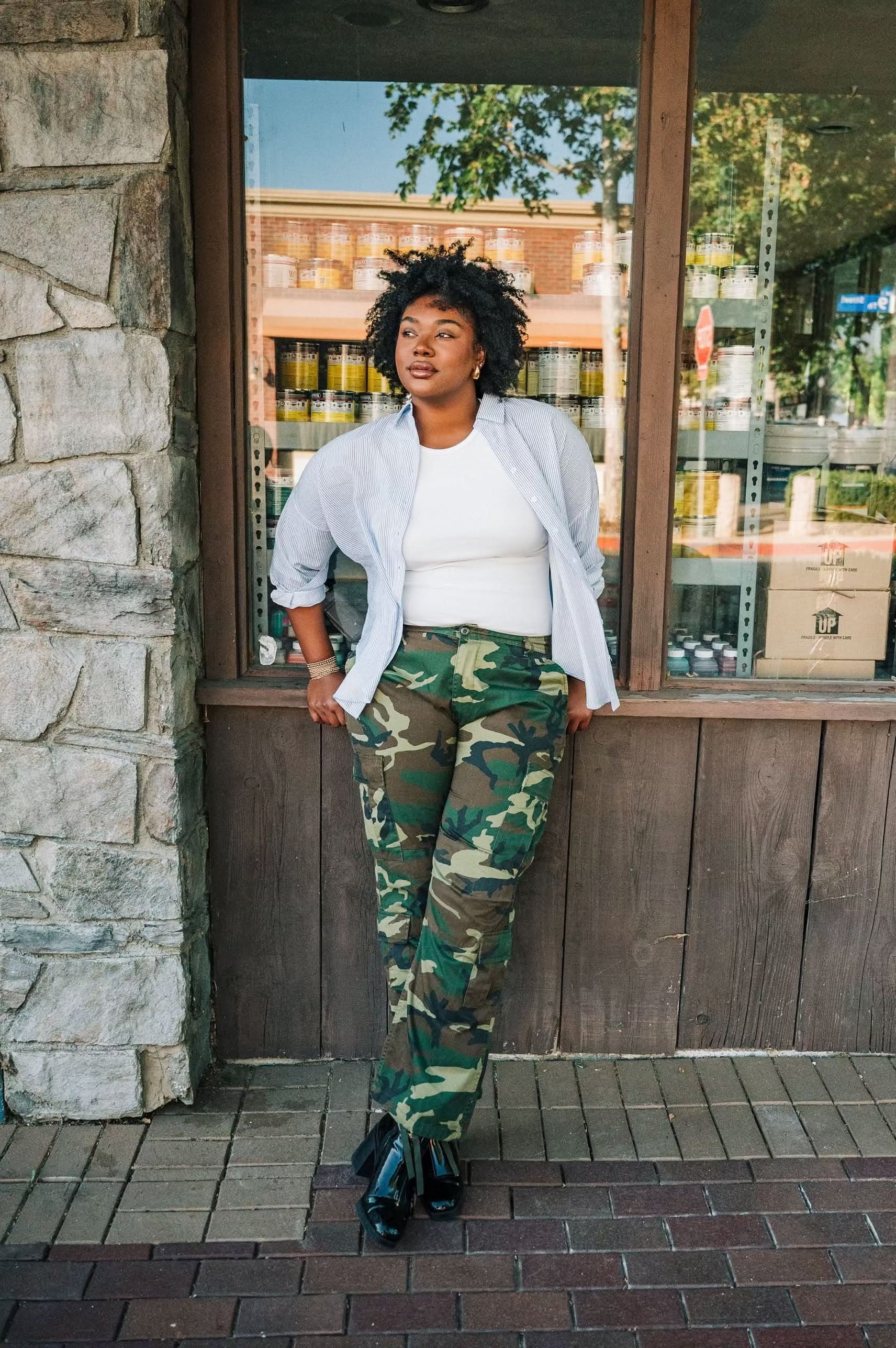 Woman wearing a white ribbed high neck tank top with a white and blue vertical button down with camo pants.