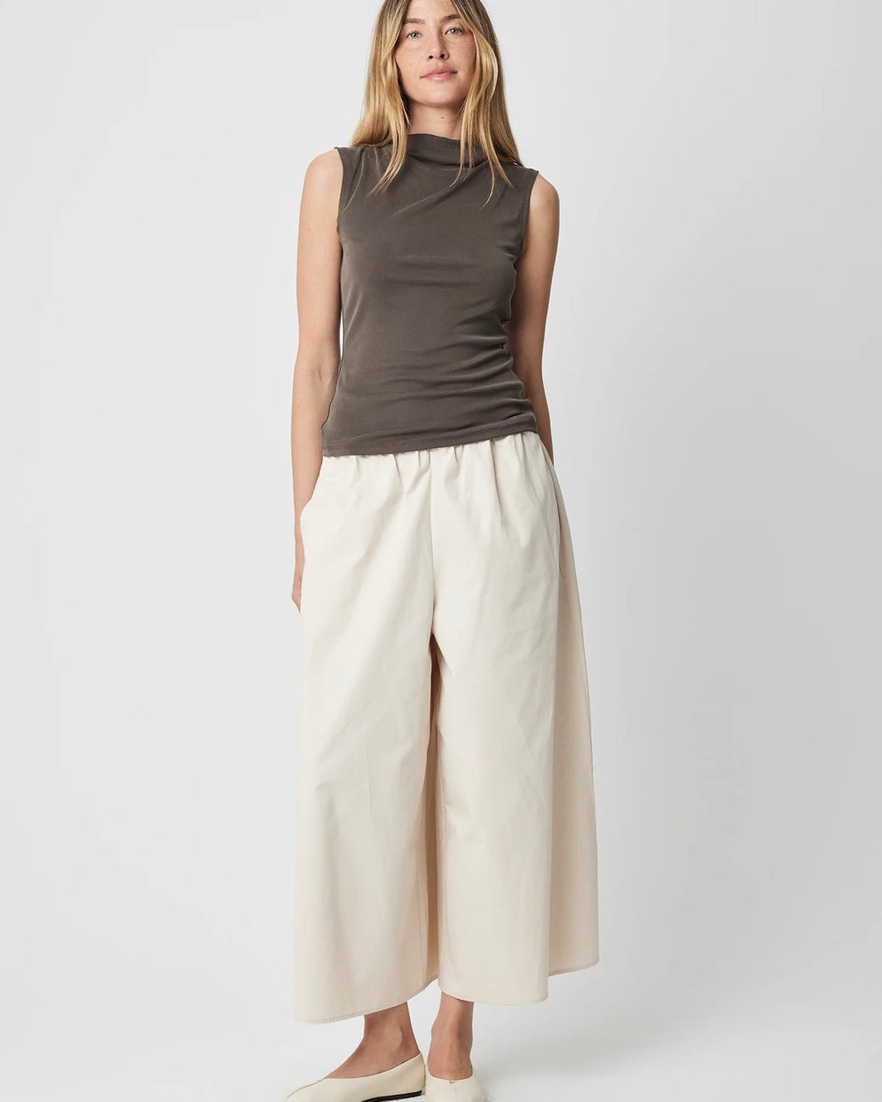 A woman modeling a sleeveless dark brown mock-neck top and cream wide-leg cropped pants with cream flats, shot against a light gray studio background.