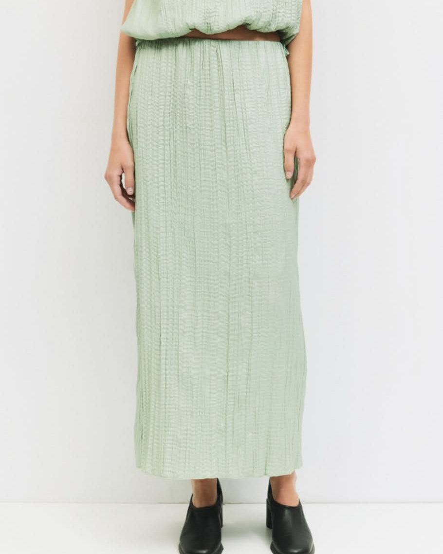 Woman standing against a white wall wearing matching sea foam, Sage green textured skirt and blouse