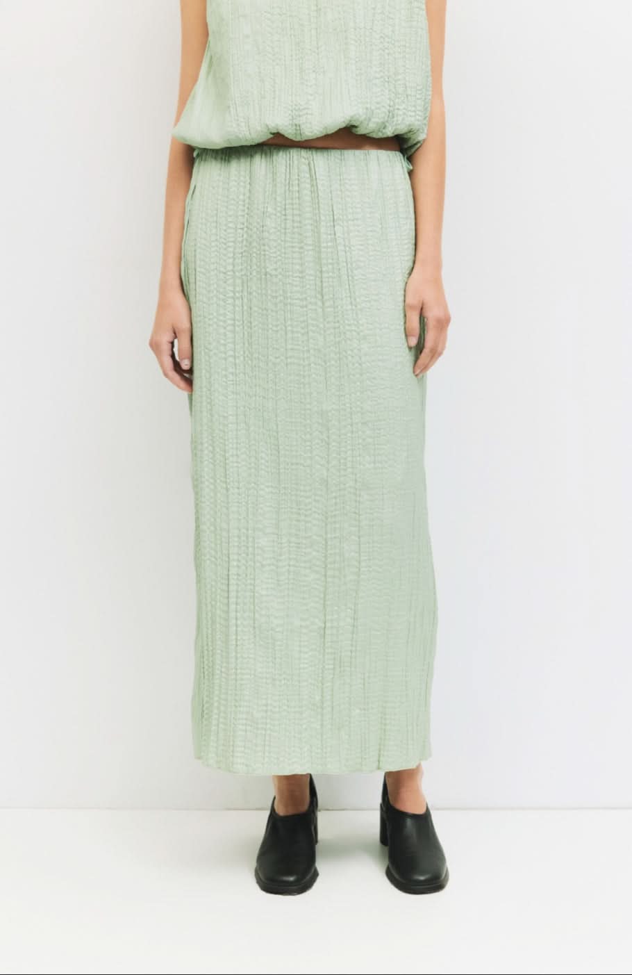 Woman standing against a white wall wearing matching sea foam, Sage green textured skirt and blouse