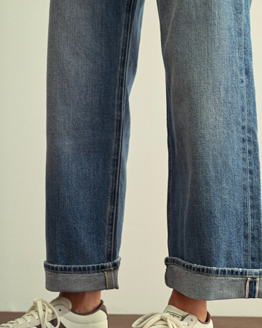 Close-up of a person wearing blue denim jeans with rolled cuffs and white sneakers against a plain light background.