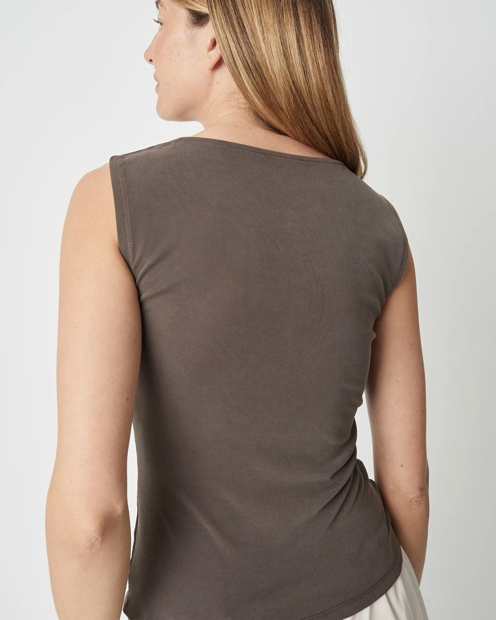 A woman shown from the back wearing a sleeveless, scoop-neck top in a muted taupe-brown color, paired with light-colored bottoms, against a plain light gray studio background.