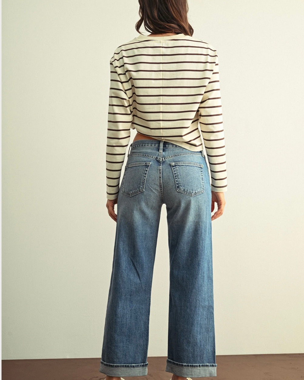 A woman viewed from behind wearing a cream and black striped long-sleeve crop top, wide-leg blue jeans, and white lace-up shoes, standing against a plain light beige wall.