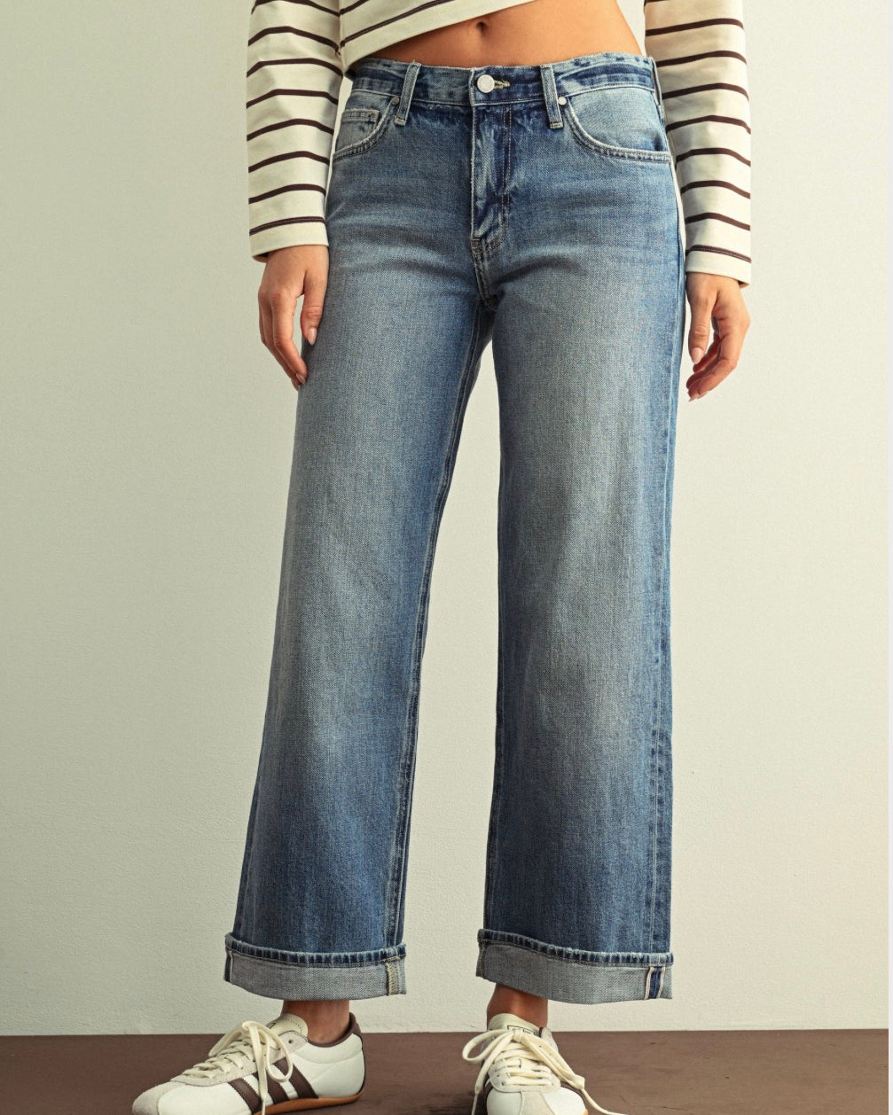 A person wearing light-wash blue wide-leg cropped jeans with rolled cuffs, paired with a cream and brown striped long-sleeve crop top and white sneakers, standing against a plain light wall.
