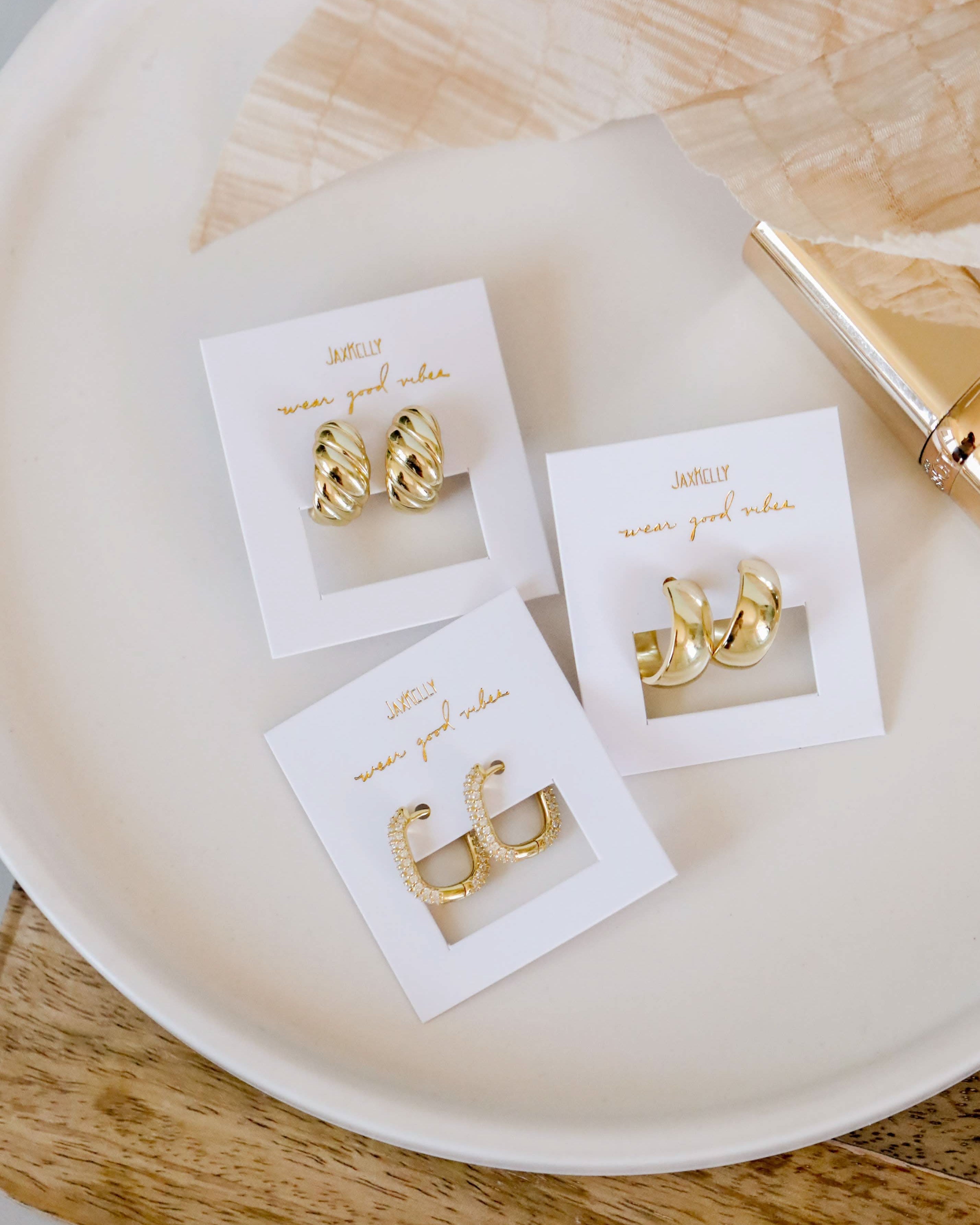 Gold hoop earrings on white display cards with 'now you know' branding, placed on a beige plate.