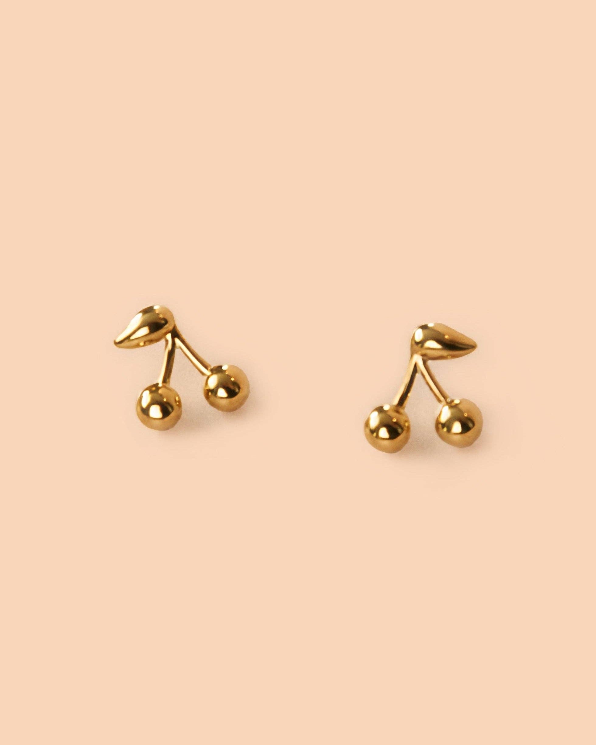Maraschino Studs Gold cherry-shaped earrings on a beige background