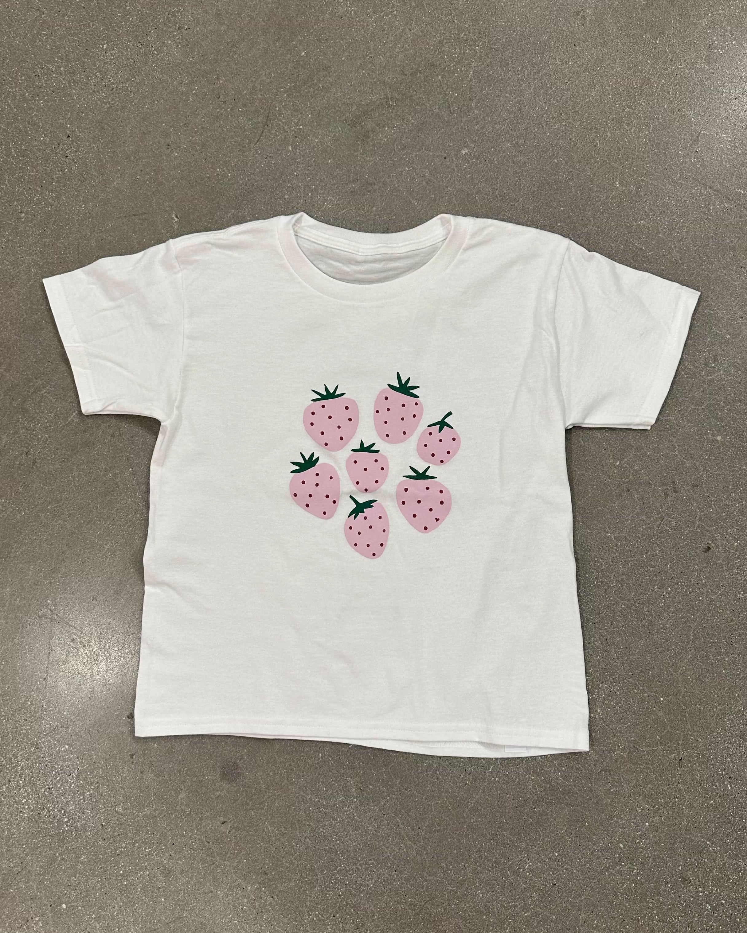 White tee-shirt with six pink strawberries on the front in front of a blank background.