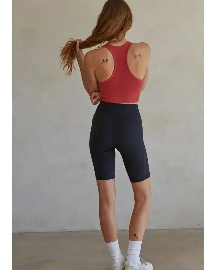 Woman wearing a red knit seamless ribbed scoop with a U-V neckline tank top with black biker shorts standing in front of a blank background.