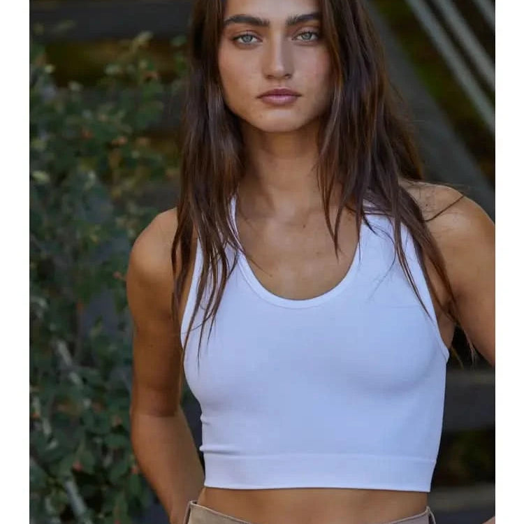 Woman wearing a white knit seamless ribbed scoop with a U-V neckline tank top with brown jeans standing behind green foliage.