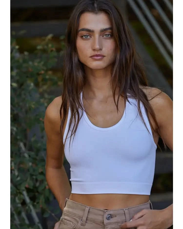 Woman wearing a white knit seamless ribbed scoop with a U-V neckline tank top with brown jeans standing behind green foliage.