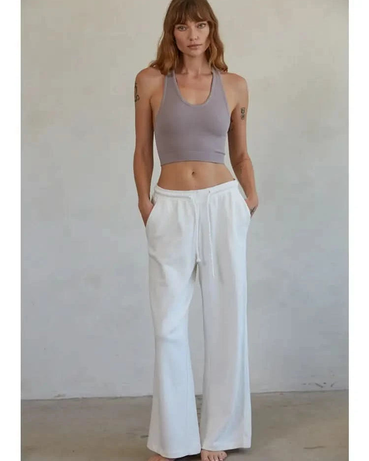 Woman wearing a mauve knit seamless ribbed scoop with a U-V neckline tank top with white drawstring sweat pants standing in front of a blank background.