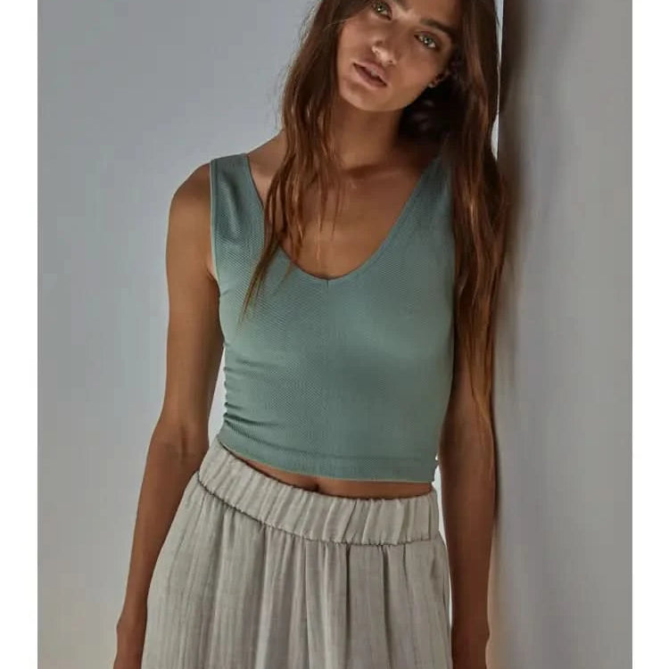 Woman wearing a light green knit seamless chevron design with a sleeveless U-V neckline tank top and white pants behind a white wall.