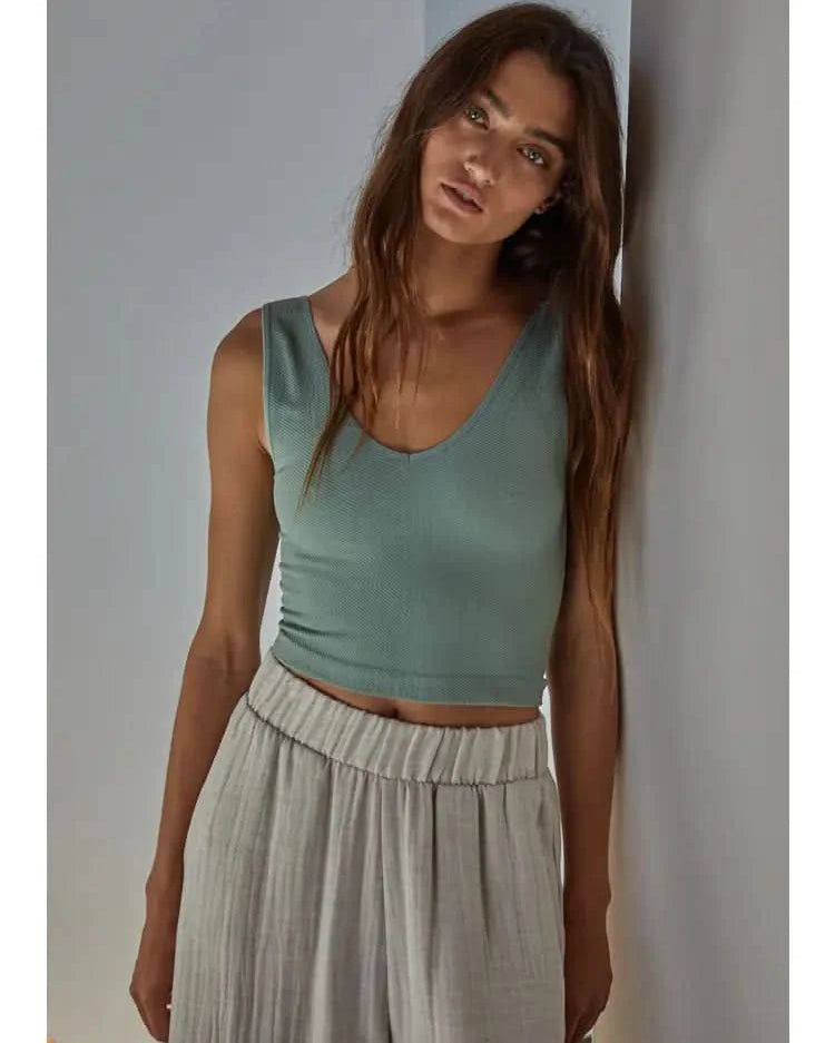 Woman wearing a light green knit seamless chevron design with a sleeveless U-V neckline tank top and white pants behind a white wall.