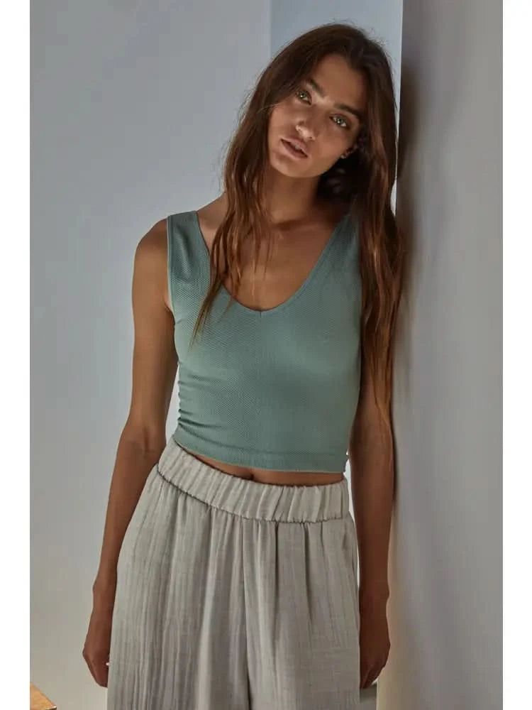 Woman wearing a light green knit seamless chevron design with a sleeveless U-V neckline tank top and white pants behind a white wall.