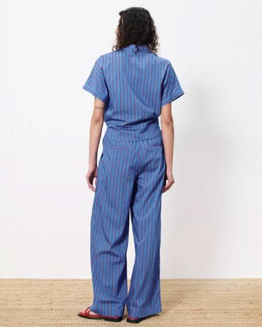 Woman facing away from viewer wearing blue botton up blouse with vertical red stripes and a front pocket. Wearing pants with the same pattern.