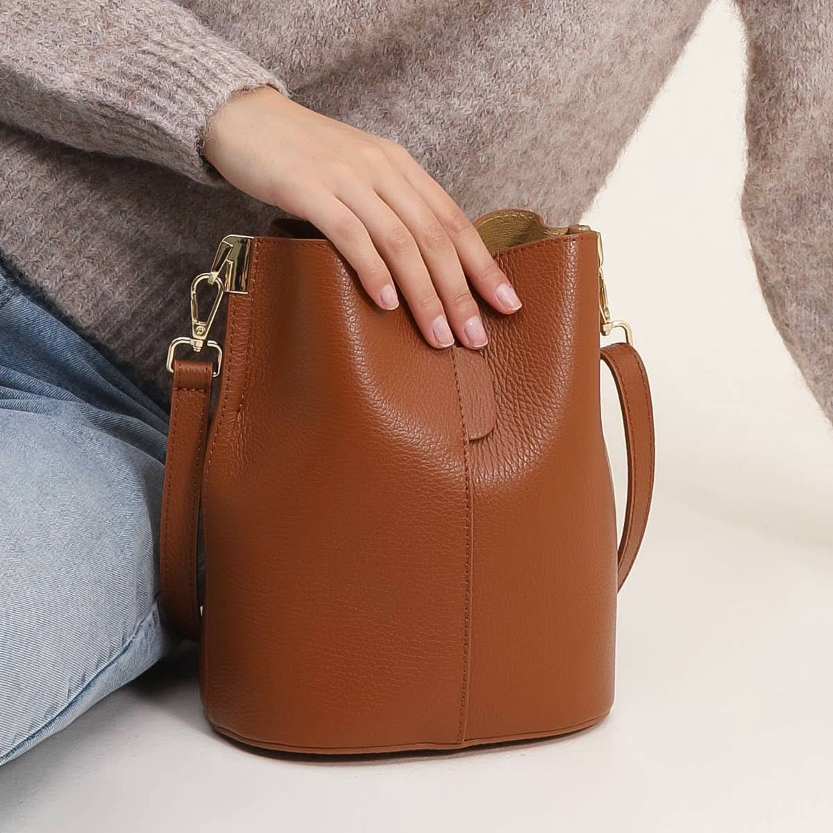 Brown leather handbag held by a person wearing a patterned sweater and jeans on a light background