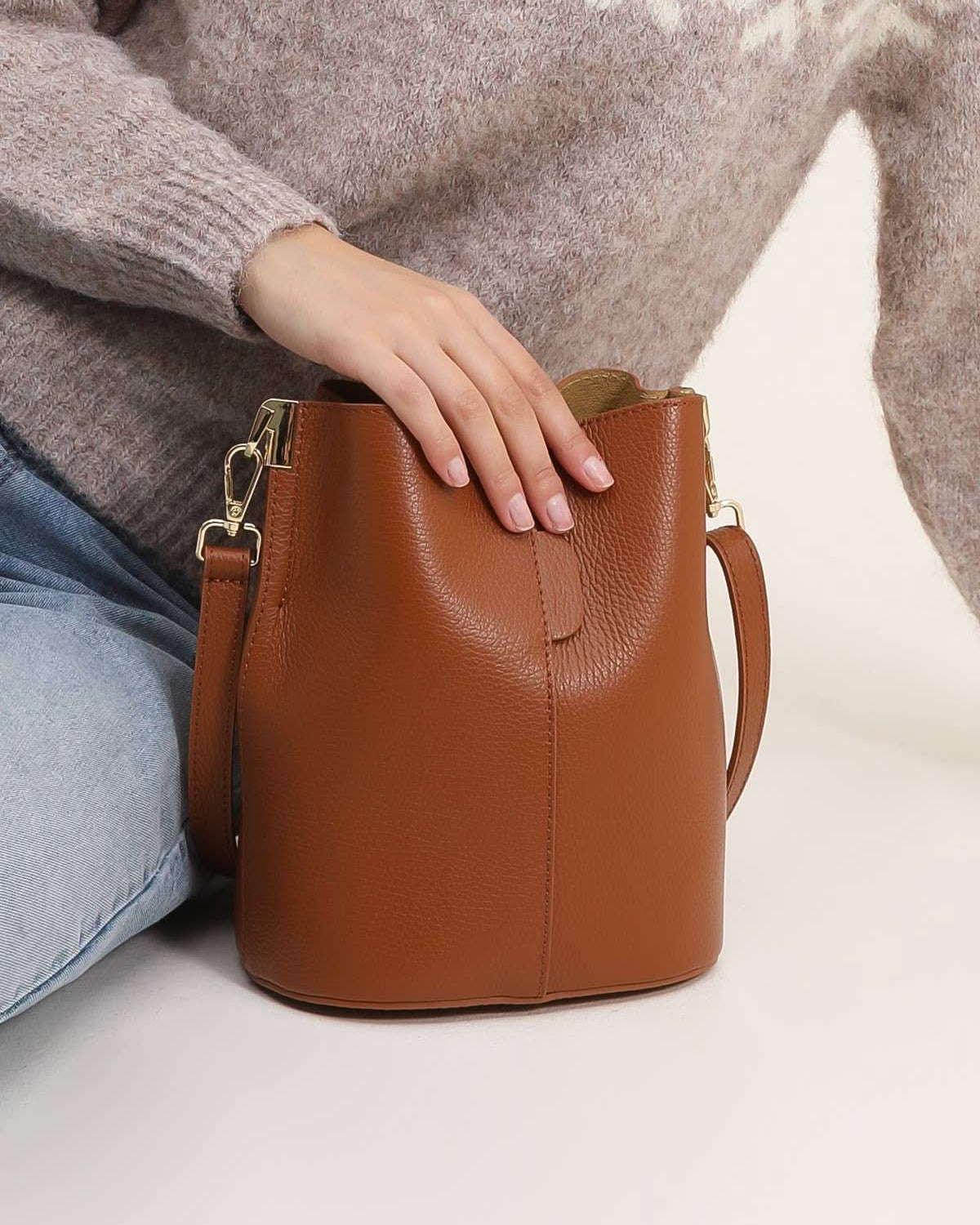Brown leather handbag held by a person wearing a patterned sweater and jeans on a light background