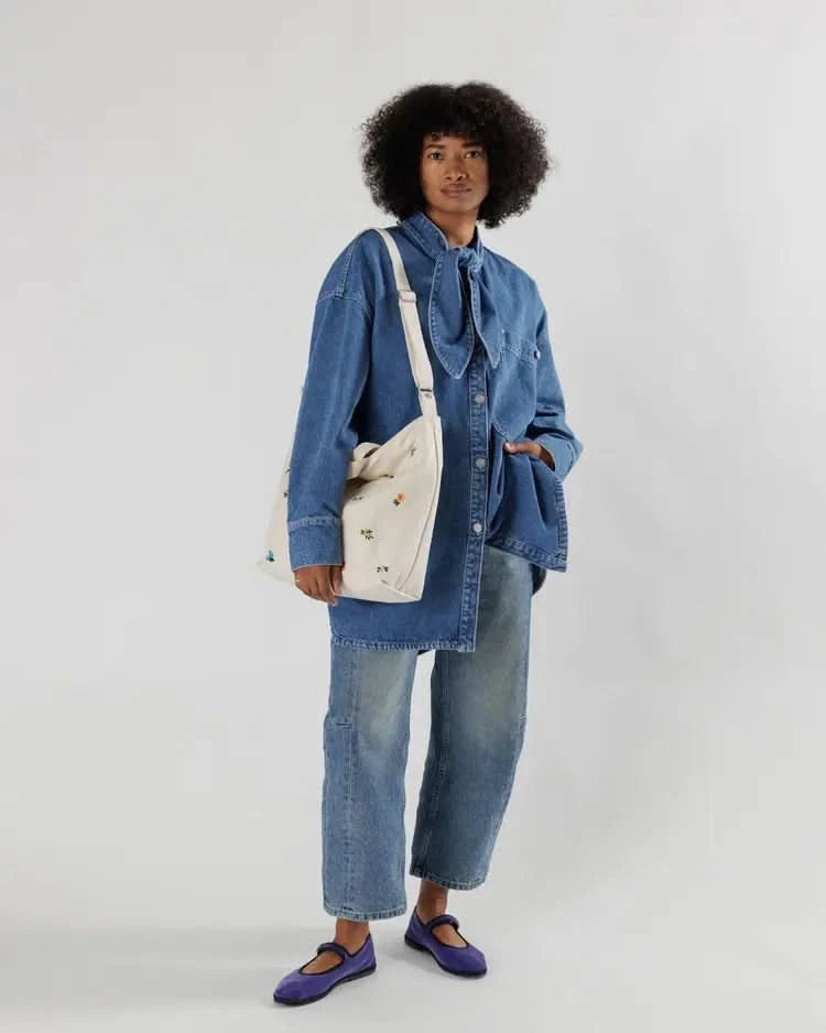 Woman in a blue outfit wearing a white over the shoulder tote bag with flowers behind a white background.
