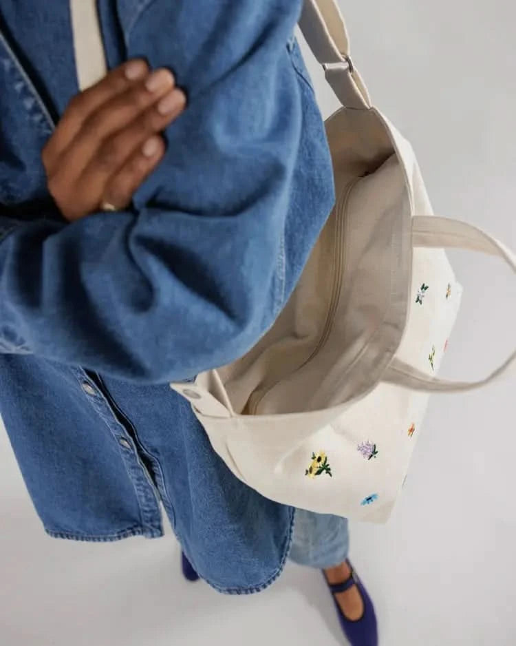 Woman wearing an over the shoulder white tote bag with flowers and a zipper behind a white background.