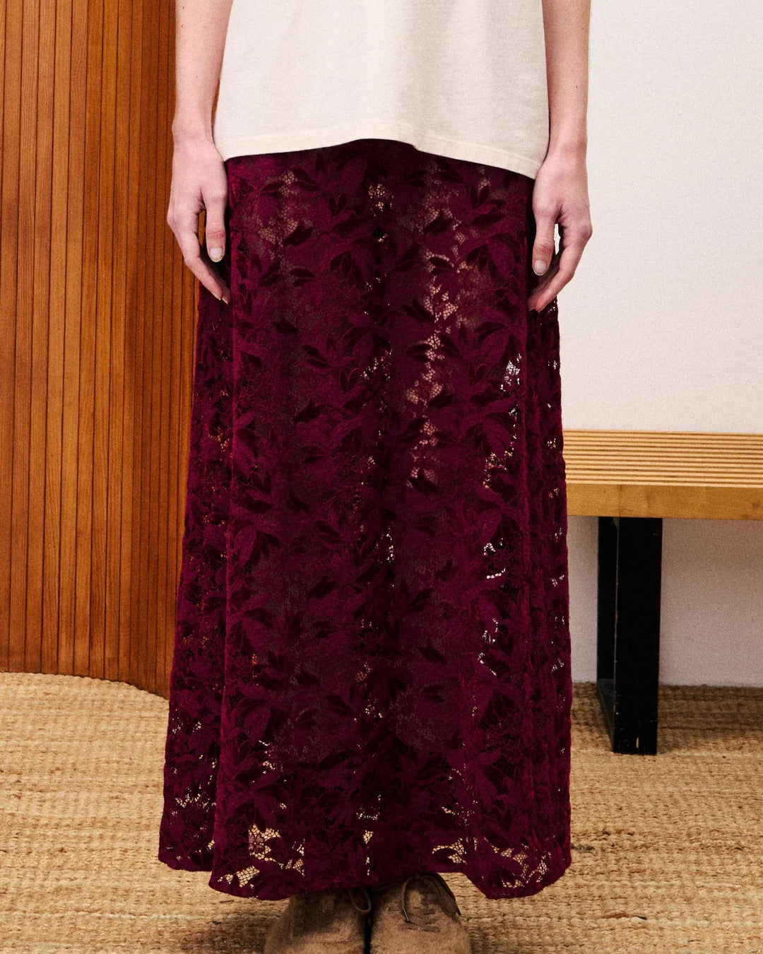 Person wearing a burgundy lace skirt with a white top and brown shoes indoors.