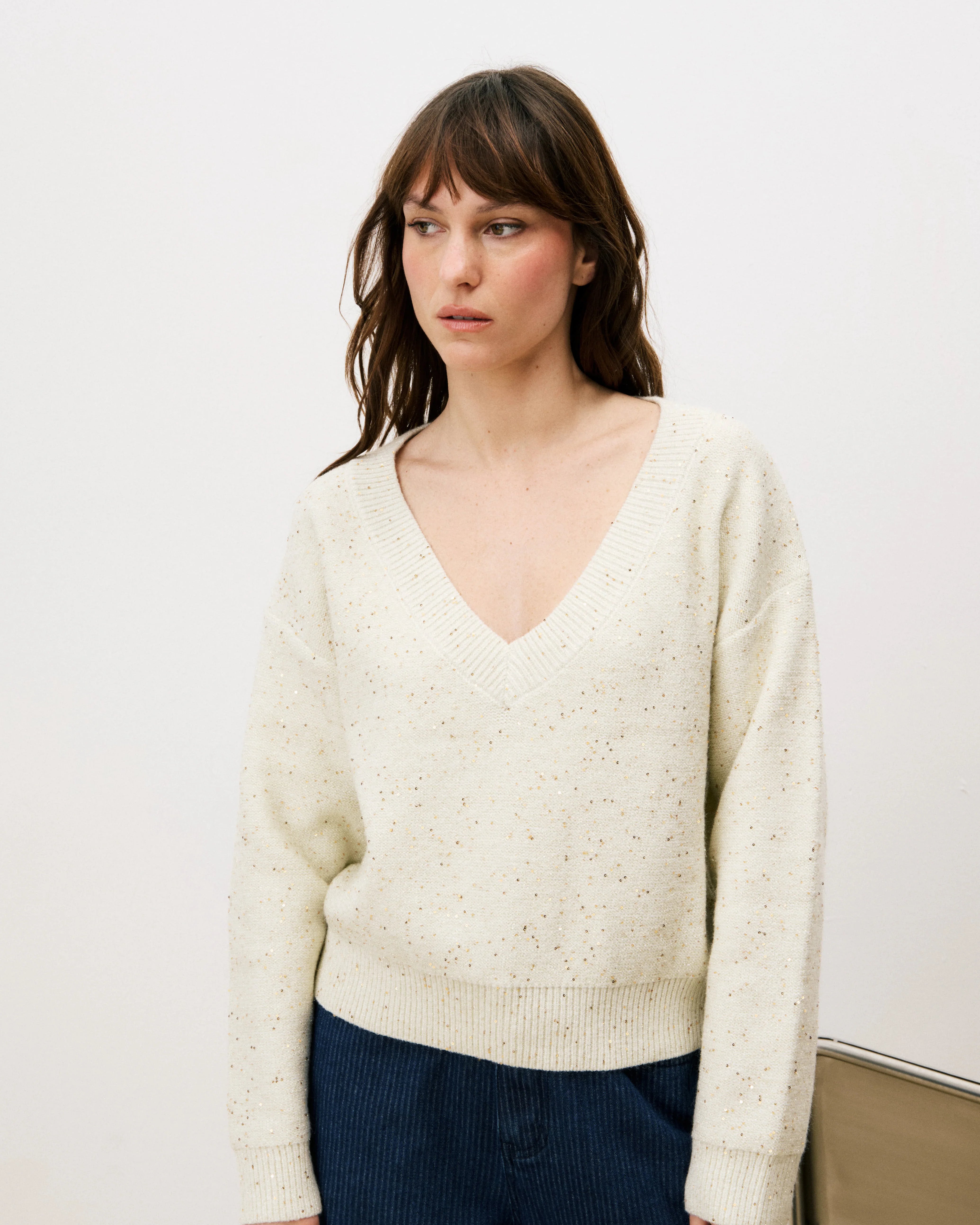 Woman wearing a cream sweater and dark pants standing against a white background