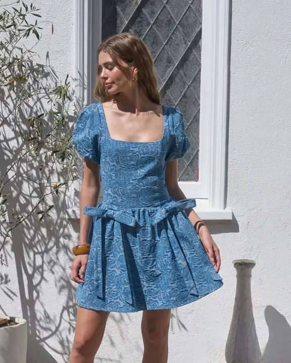 Woman wearing a blue dress standing outdoors against a white wall.