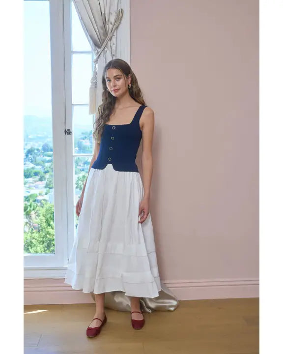 Woman wearing a blue top and white skirt standing in a room with large windows.