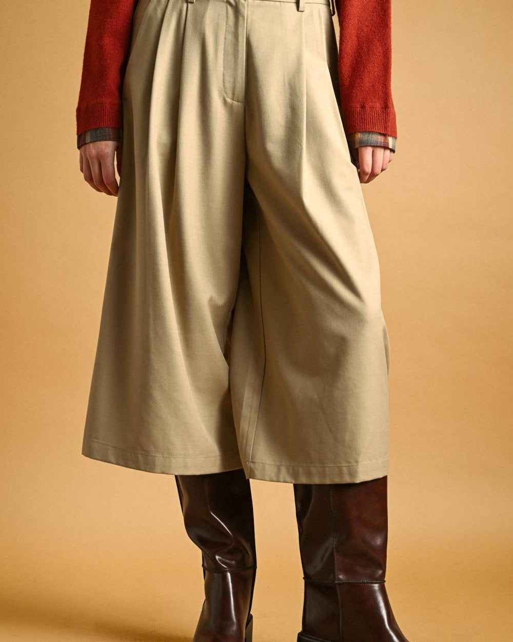 A person wearing wide-leg cropped beige pants, a rust red cardigan, and dark brown leather knee-high boots against a warm tan studio background.