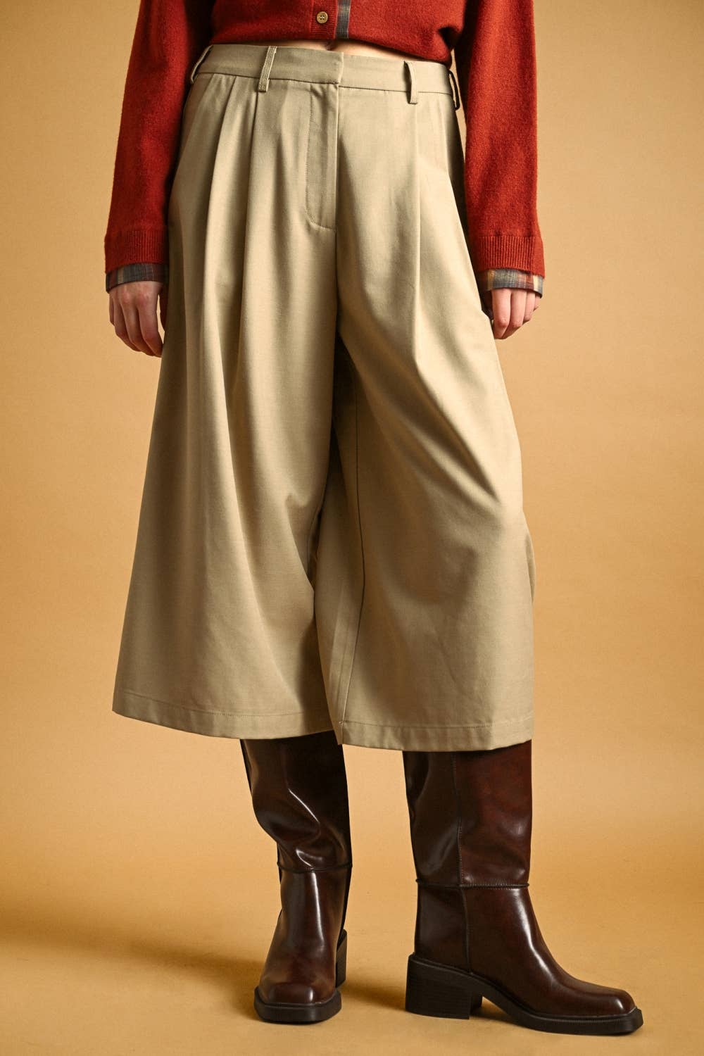 A person wearing wide-leg cropped beige pants, a rust red cardigan, and dark brown leather knee-high boots against a warm tan studio background.