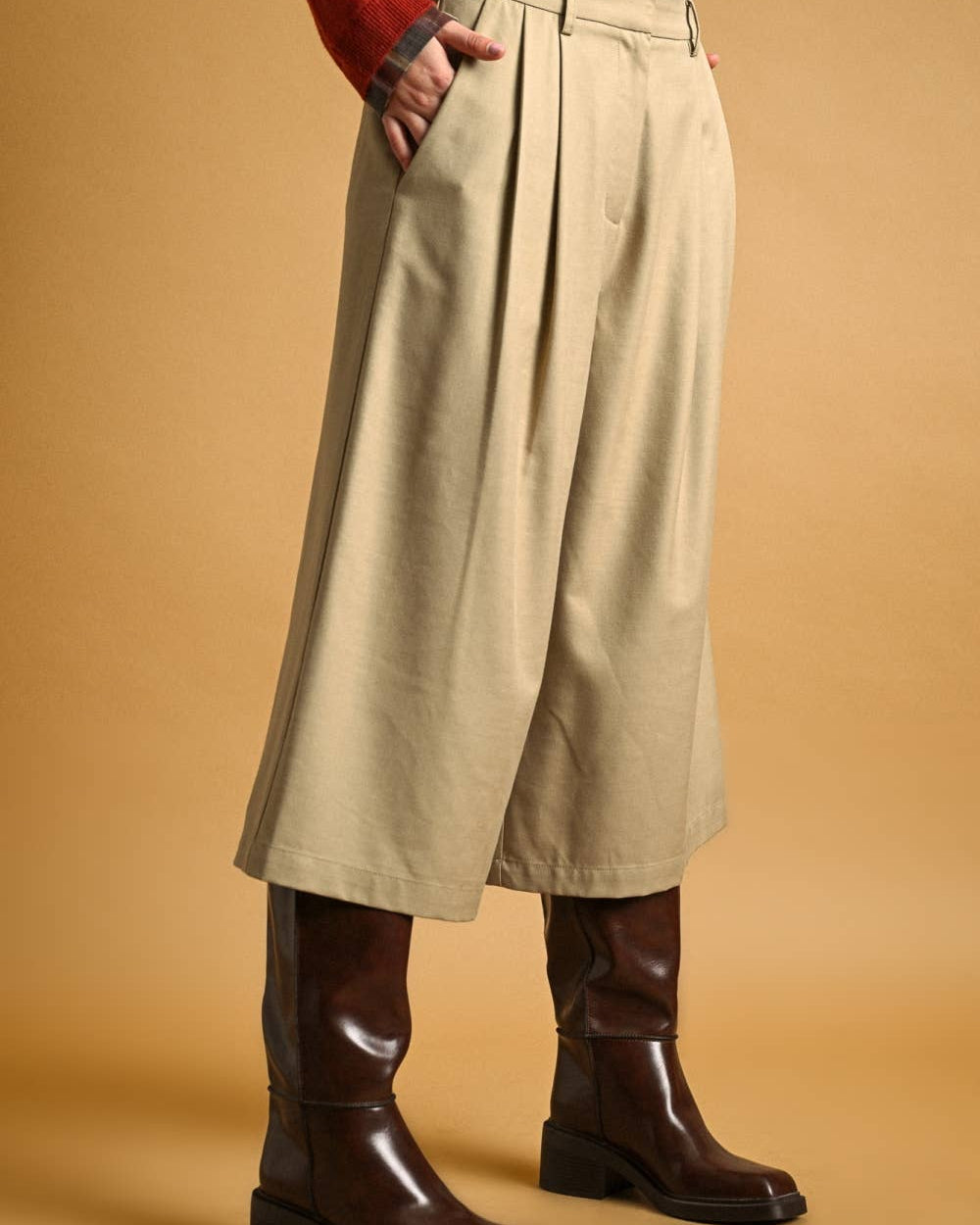 A model is shown from the waist down wearing high-waisted, wide-leg pleated cropped trousers in a light khaki-beige color, styled with a red cardigan and dark brown leather boots against a solid warm tan background.
