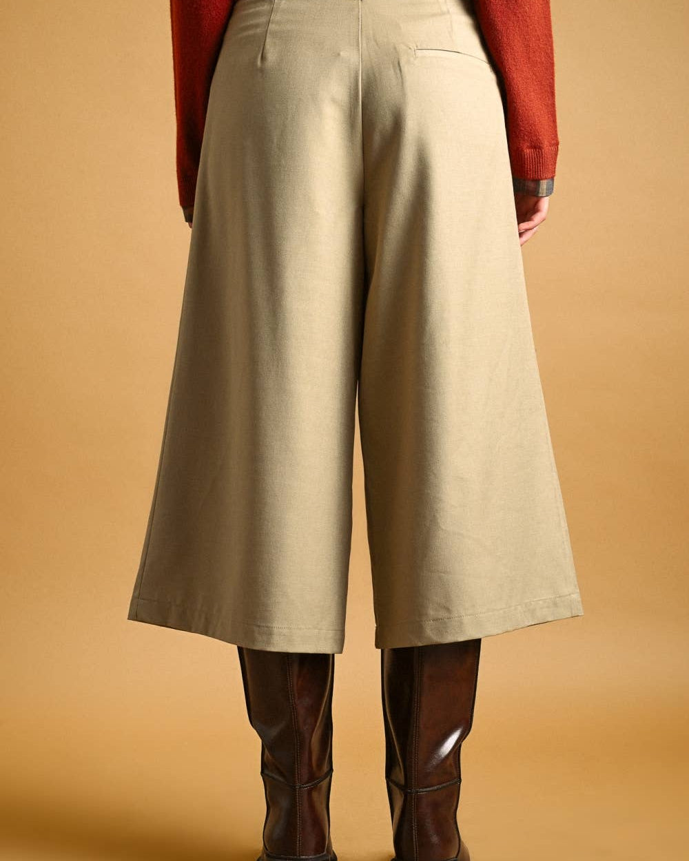 A person shown from the back wearing high-waisted, wide-leg cropped pants in a light beige/khaki color, paired with a rust-red sweater and dark brown knee-high boots, against a solid warm tan background.