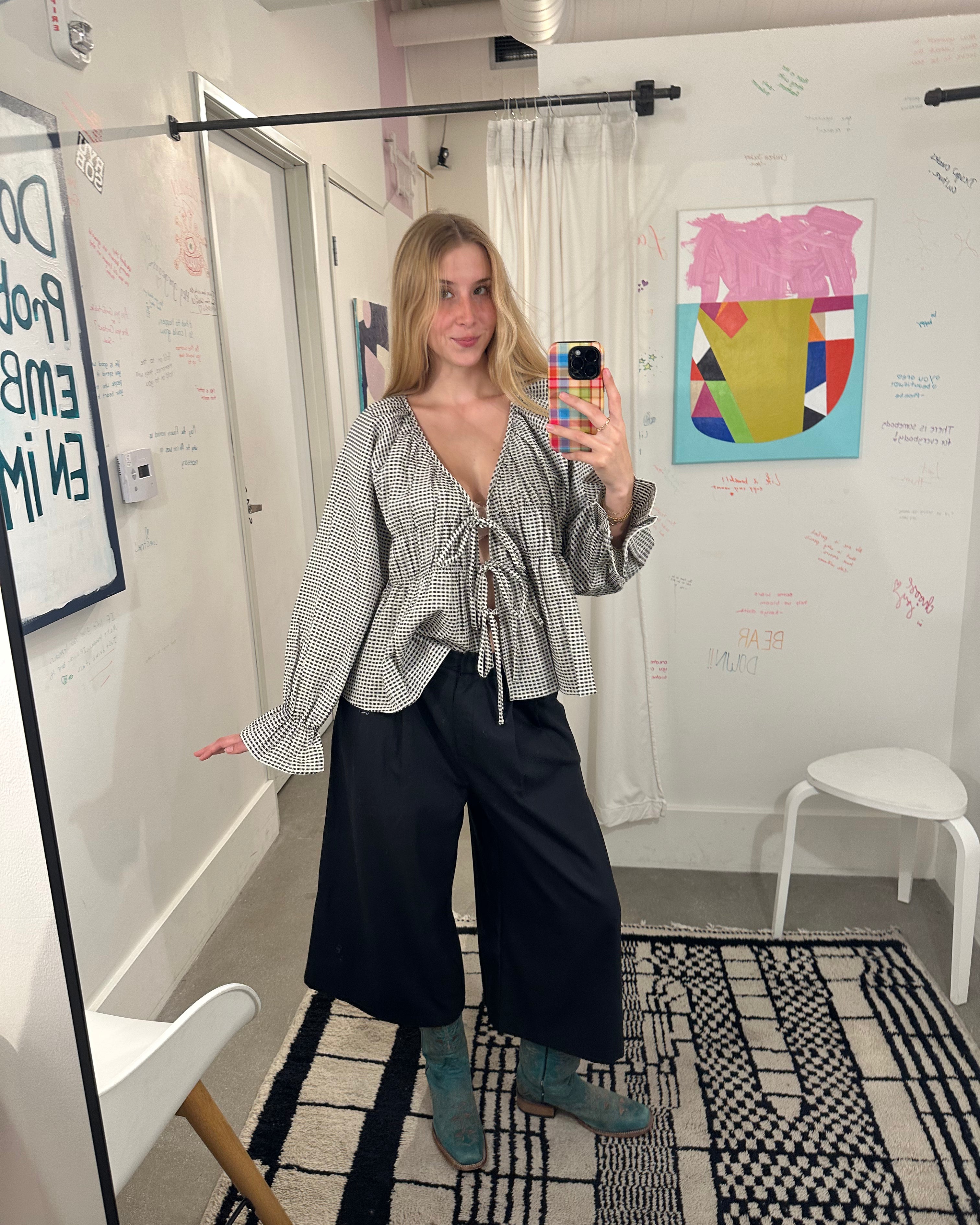 A woman takes a mirror selfie in a room, wearing a gray and white vertically striped long-sleeve blouse with a tie neckline, black wide-leg pants, and teal boots.