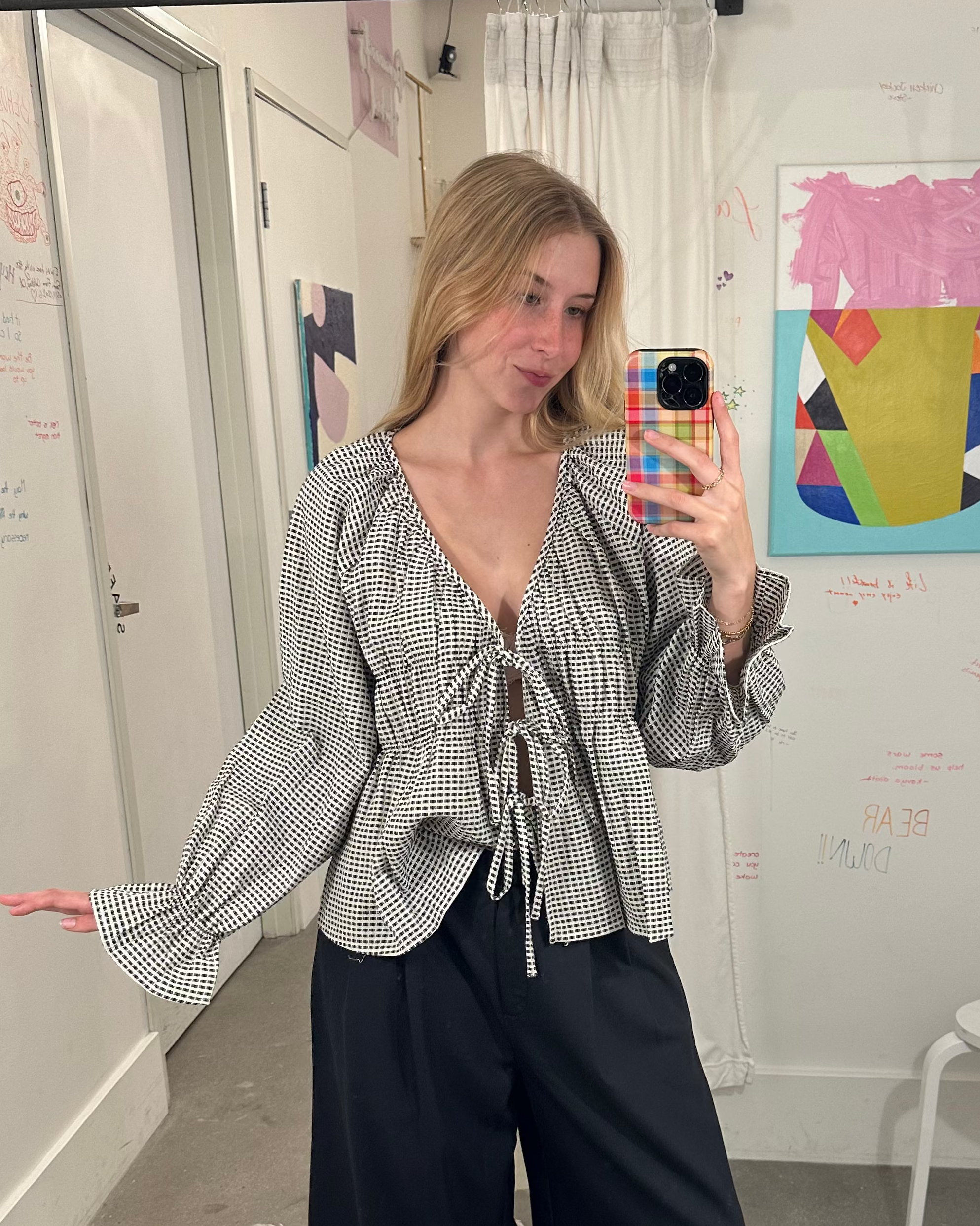 A woman takes a mirror selfie in a fitting room wearing a black and white vertically striped long-sleeve blouse with a tie-front detail and black wide-leg pants.