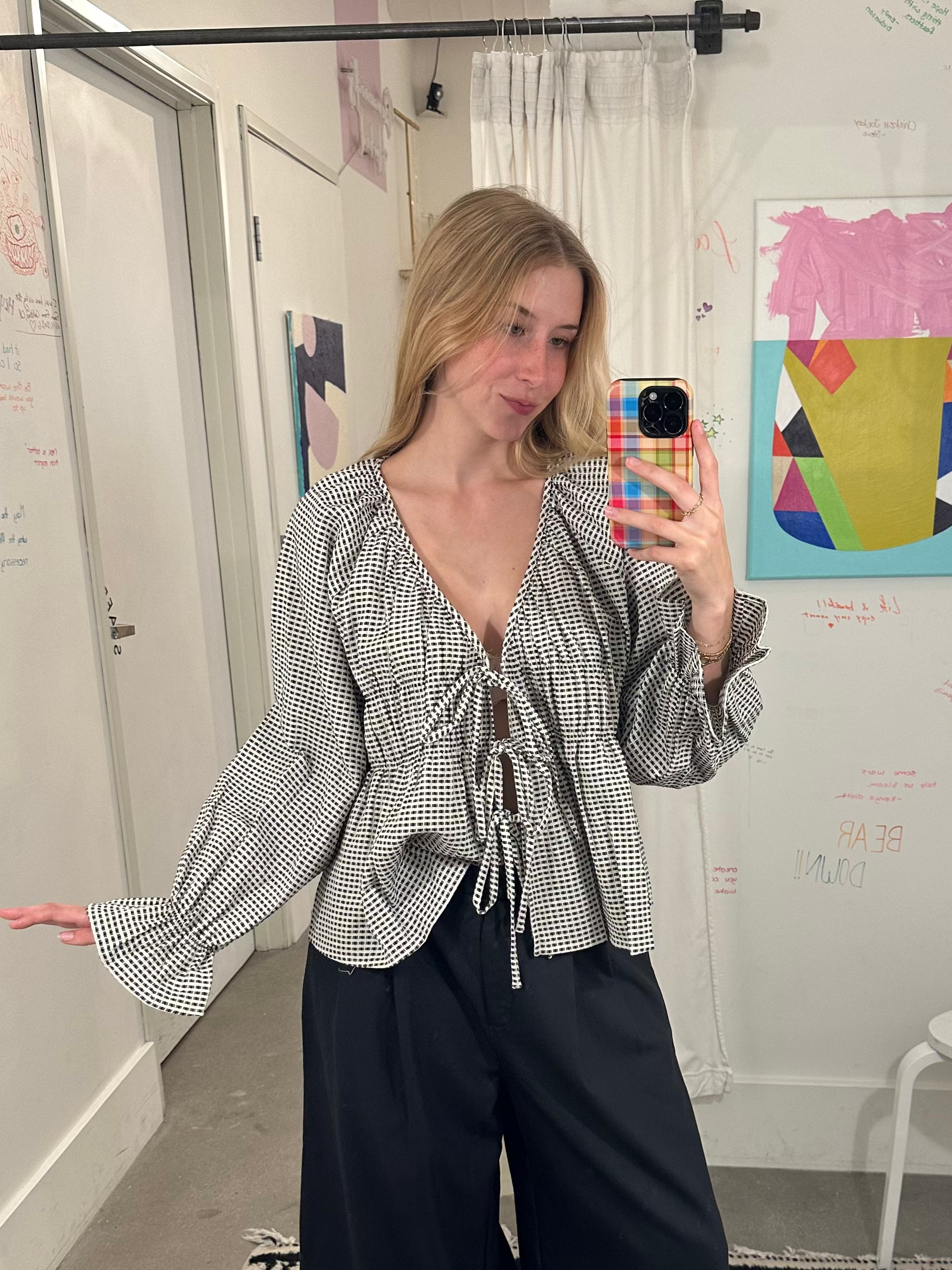 A woman takes a mirror selfie in a fitting room wearing a black and white vertically striped long-sleeve blouse with a tie-front detail and black wide-leg pants.