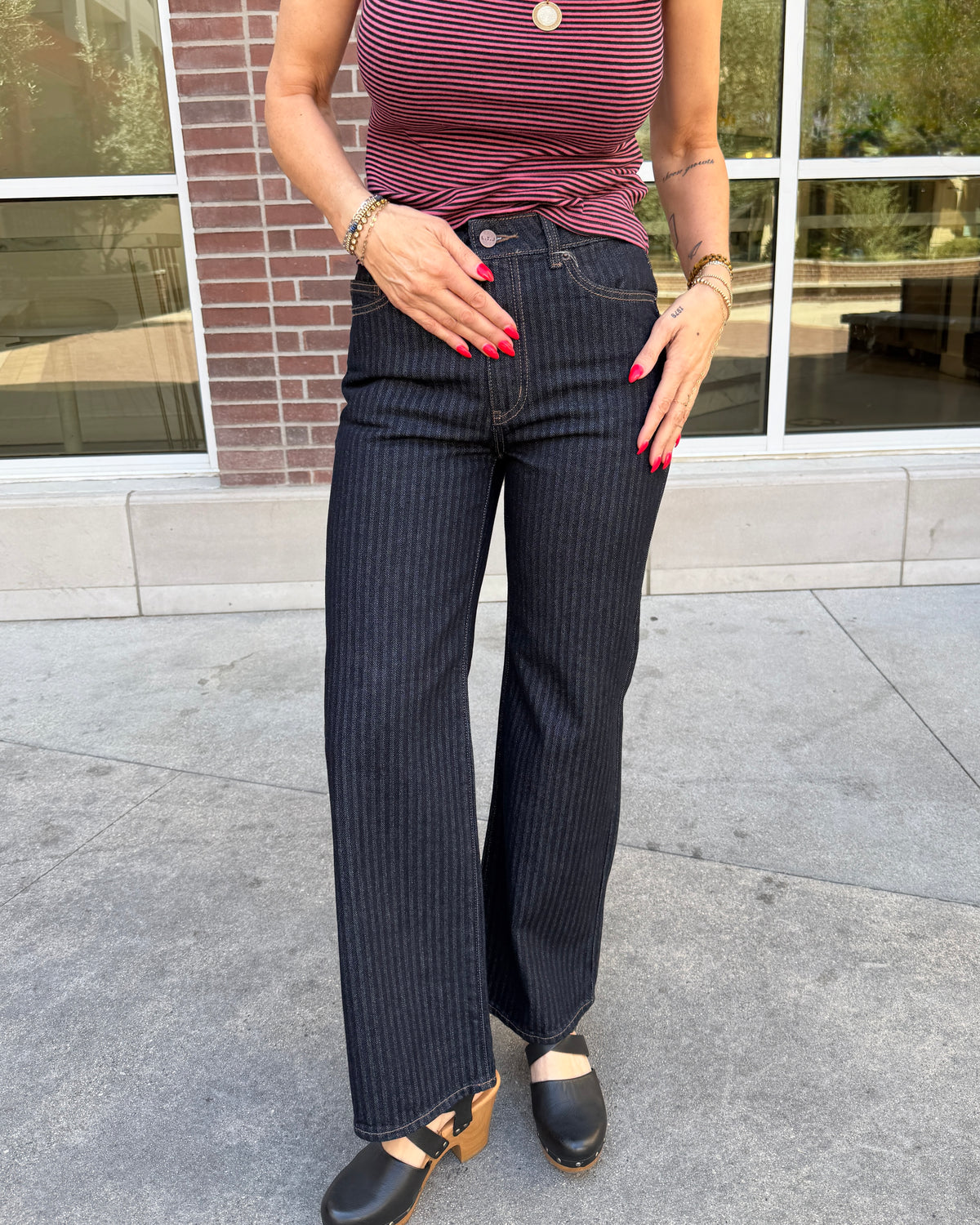 A person wearing dark navy pinstriped jeans, a burgundy and pink striped top, and black clogs, standing outdoors in front of a brick building with large windows.