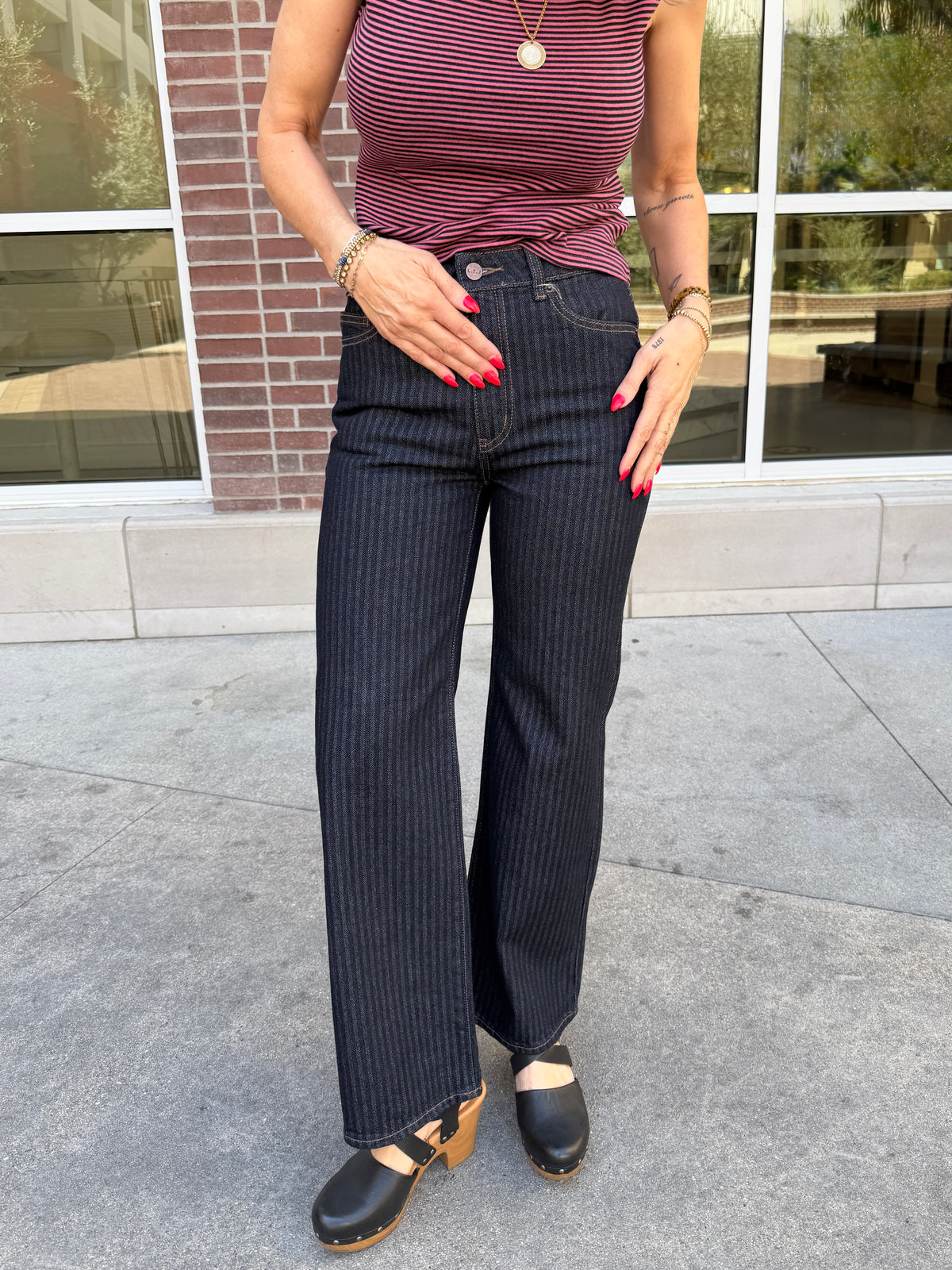 A person wearing dark navy pinstriped jeans, a burgundy and pink striped top, and black clogs, standing outdoors in front of a brick building with large windows.