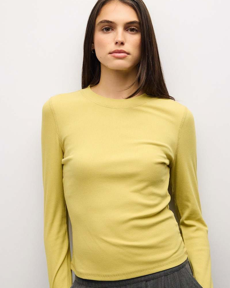 A woman wearing a fitted, long-sleeve crewneck top in a soft pastel yellow, paired with a dark gray skirt, standing against a plain light gray background.