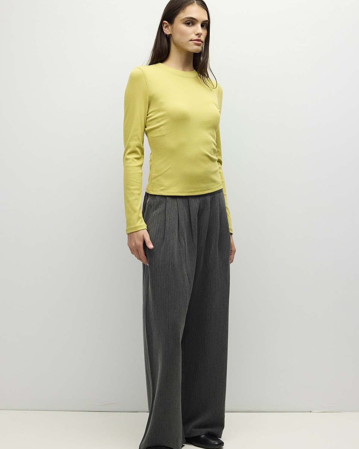 A woman modeling a fitted long-sleeve top in pale yellow-green paired with wide-leg dark gray trousers, standing against a plain light gray studio background.