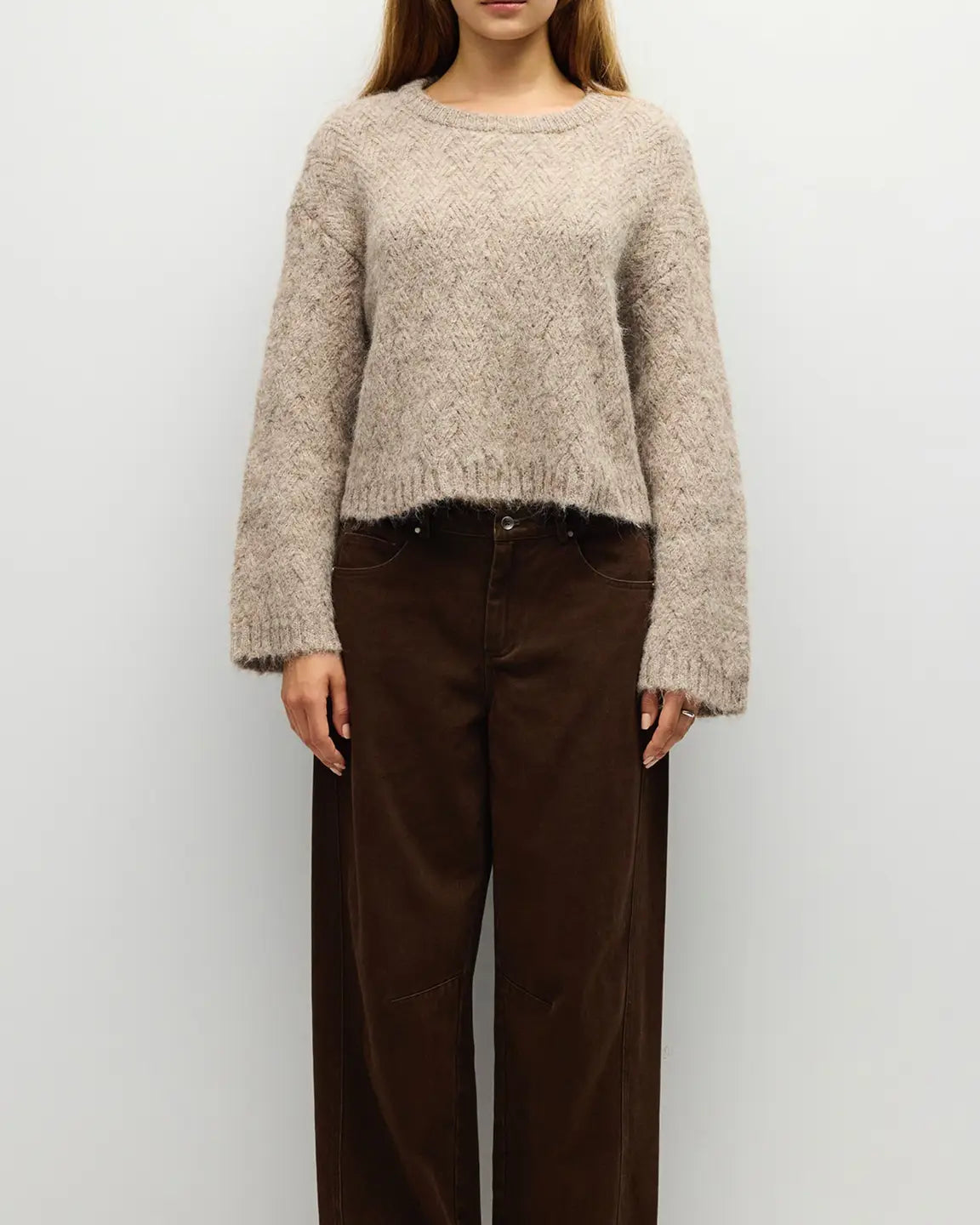 A woman wearing a fuzzy cropped knit sweater in a light taupe-beige color, paired with dark brown wide-leg pants, standing against a plain light gray background.
