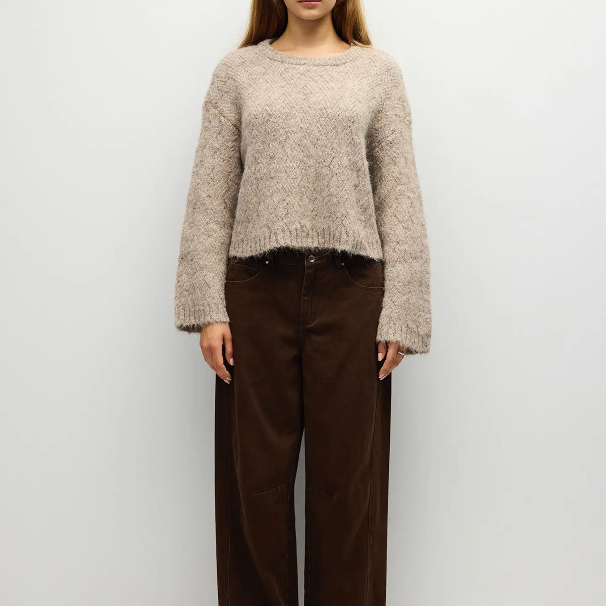 A woman wearing a fuzzy cropped knit sweater in a light taupe-beige color, paired with dark brown wide-leg pants, standing against a plain light gray background.