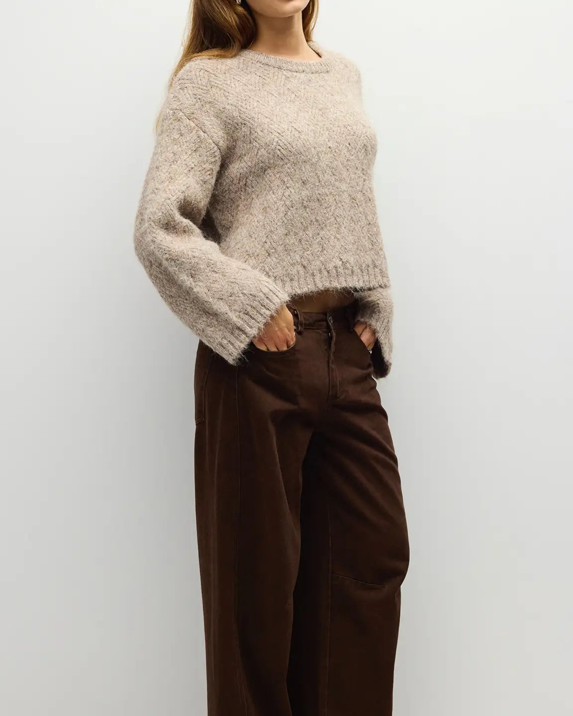 A woman wearing a chunky, fuzzy knit sweater in a light oatmeal beige color and dark brown wide-leg trousers, standing against a plain light gray wall.