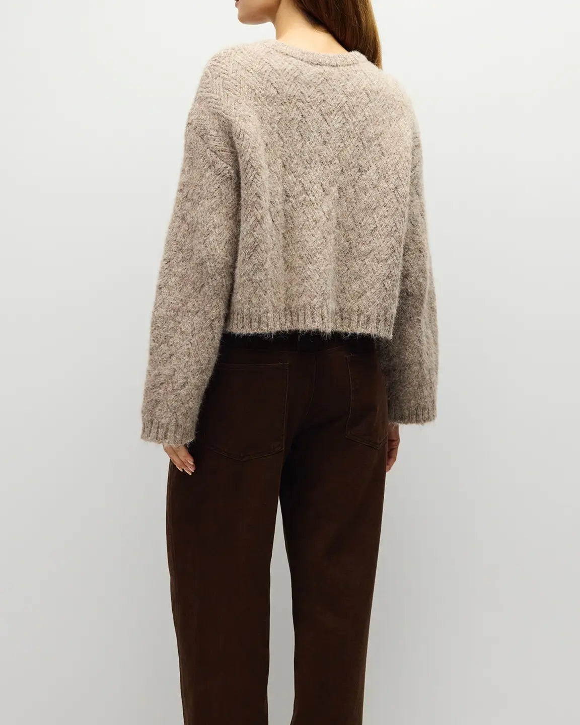 A woman shown from the back wearing a cropped, textured beige knit sweater and dark brown pants against a plain light gray background.