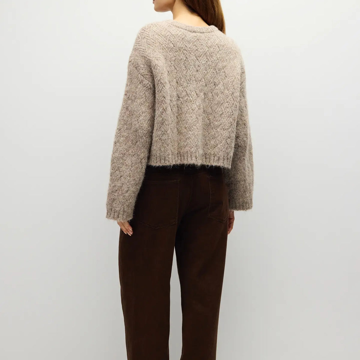 A woman shown from the back wearing a cropped, textured beige knit sweater and dark brown pants against a plain light gray background.