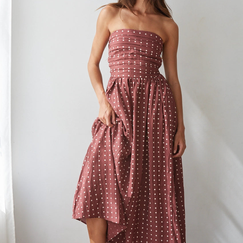 A woman models a strapless dusty rose midi dress with white polka dots, featuring a ruched bodice and a flowing skirt, paired with brown block-heel sandals against a white wall.