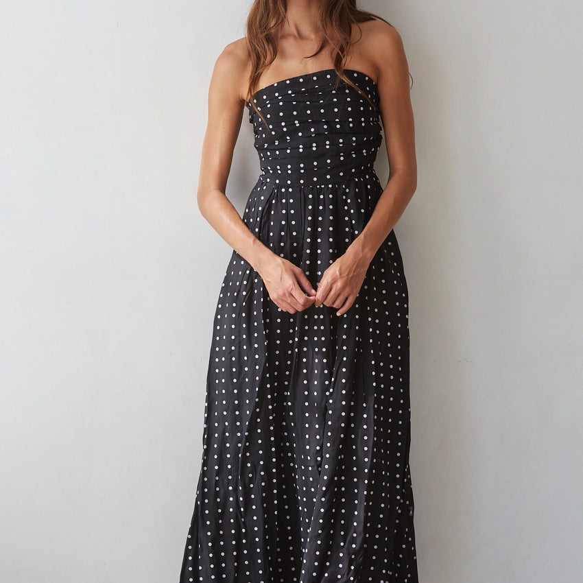 A woman modeling a strapless black midi dress with a white polka dot pattern, standing against a plain light gray wall.