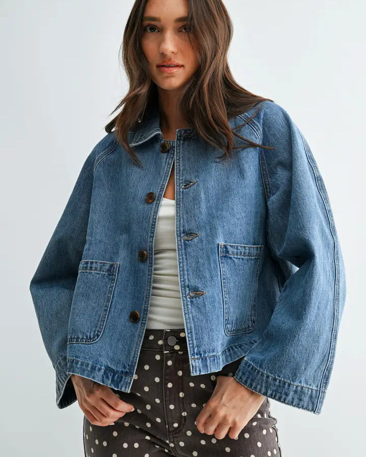 A woman wearing a cropped, oversized light blue denim jacket over a white top and dark brown polka-dot pants, photographed against a plain light gray studio background.
