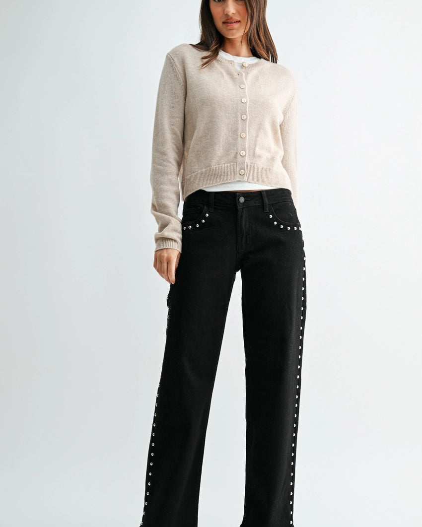 woman wearing black jeans with silver studs and a cream sweater in front of a white background.